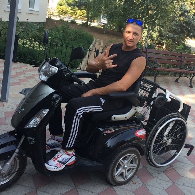 На фото: Алексей Метла - молодой мужчина в спортивных штанах и черной футболке - сидит на трехколесном мопеде. К задней части мопеда прикреплена инвалидная коляска