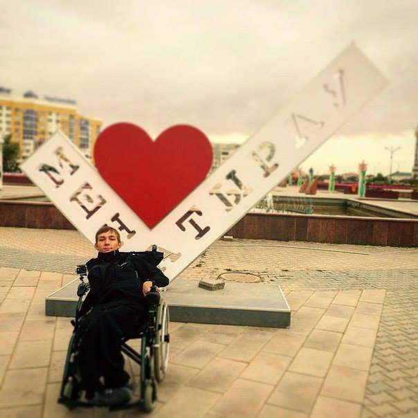 На фото: Артур Руссман в коляске фотографируется рядом с большим знаком в виде сердца с буквами: Атырау
