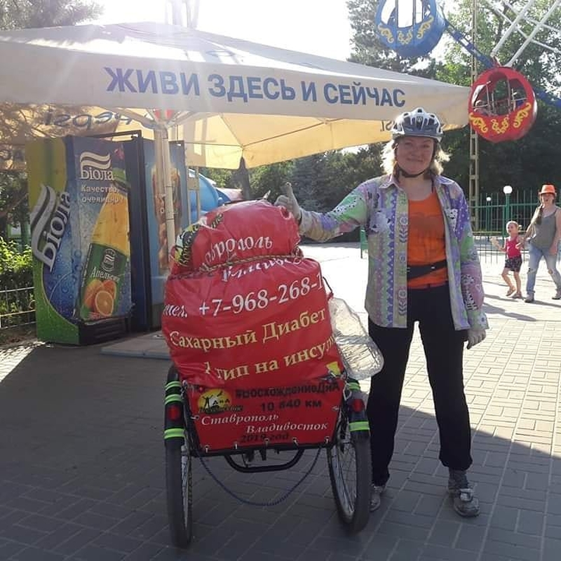На фото: Наталья с велосипедом стоит под тентом с надписью: "Живи здесь и сейчас"