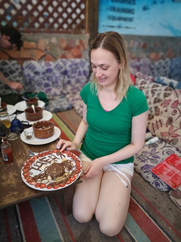 На фото: стоянка в пустыне. Евгения Малышко в зеленой футболке и шортах сидит на ковре в лагере бедуинов. Позади нее - мягкие подушки. Перед девушкой стоит низкий столик с тарелками и едой. Перед Евгенией на большом блюде импровизированный торт - бананы, клубника, залитые шоколадом. На тарелке надпись шоколадными буквами: Женя