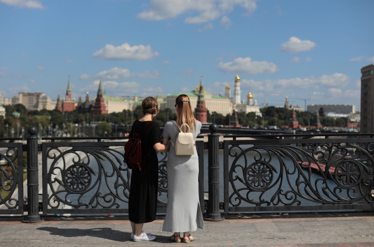 На фото: Солнечный летний день. Евгения Малышко в длинном светлом платье и в темных очках проводит экскурсию для девушки в черном платье с завязанными глазами. Фотография сделана на мосту над рекой, с видом на Кремль