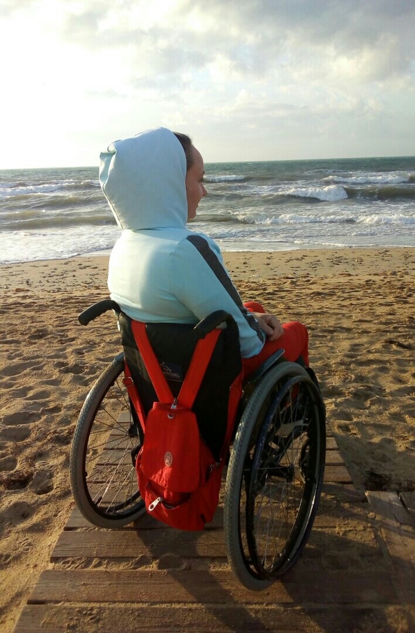 На фото: Елена Новикова в куртке с капюшоном сидит в инвалидной коляске на берегу моря