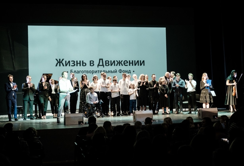 На фото: в темном зале на освещенной сцене в ряд выстроились взрослые и дети - участники мероприятия. Некоторые ребята с протезами, другие на колясках. Позади них на большом экране написано название фонда "Жизнь в Движении"