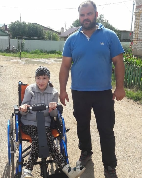 На фото: Кирилл Климов в голубой футболке и черных брюках стоит рядом с девочкой, сидящей в инвалидной коляске с электроприставкой