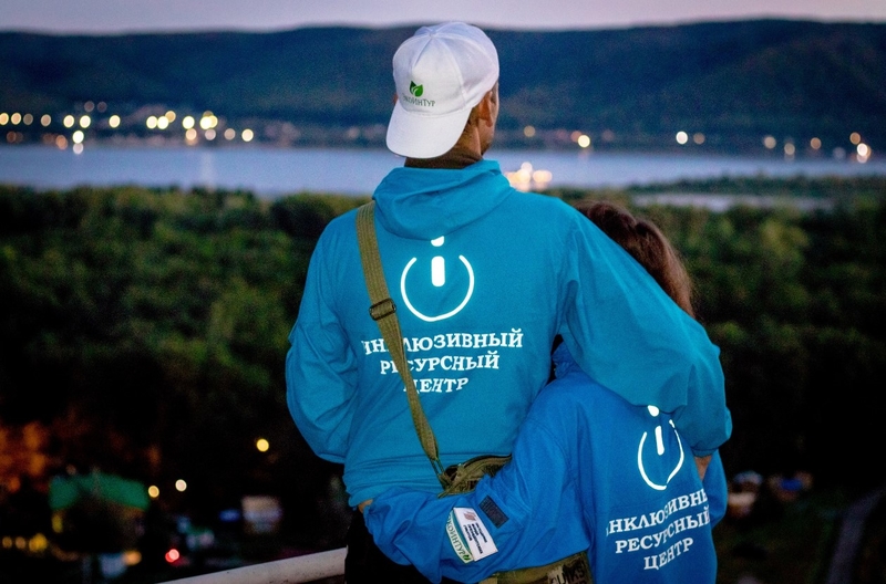 На фото: парень и девушка в ветровках с надписью "Инклюзивный ресурсный центр" стоят и любуются на вечерний лагерь и реку