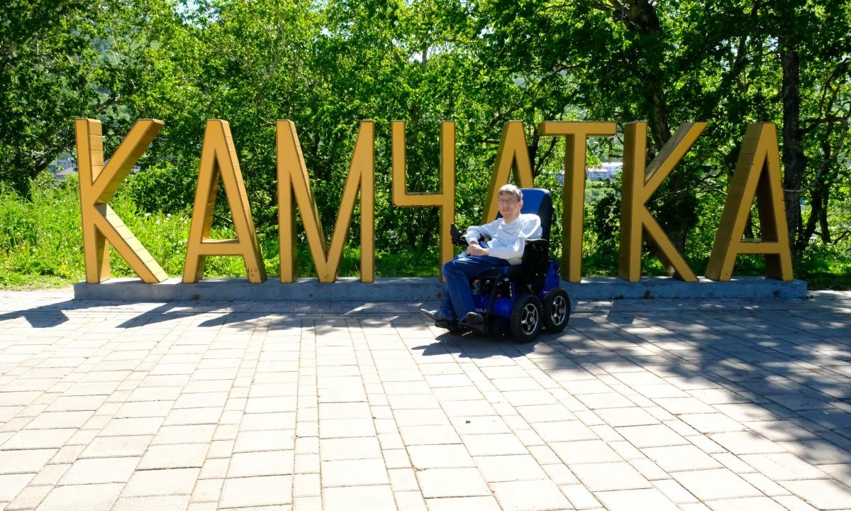 На фото: Максим Аркуша - невысокий мужчина в электроколяске, фотографируется перед арт-объектом - надписью Камчатка большими желтыми буквами в человеческий рост на Никольской сопке в Петропавловске-Камчатском