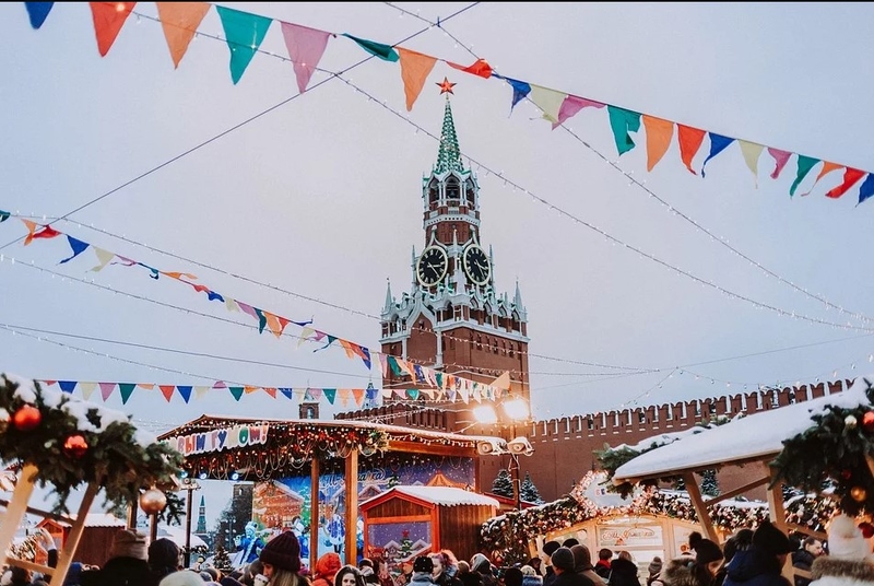 На фото: Красная площадь в Москве, празднично украшенная к Новому году. Над площадью висят разноцветные флажки, люди ходят между деревянными домиками, где продают игрушки, конфеты и прочие новогодние сувениры