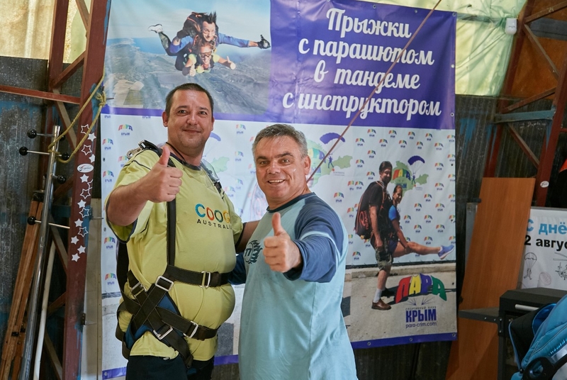 На фото: участник проекта в экипировке для прыжка стоит рядом с инструктором в ангаре. Оба показывают большим пальцем вверх