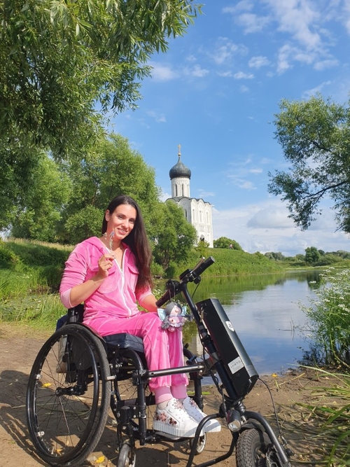 На фото: Нина Веденина-Меерсон в инвалидном кресле с приставкой на берегу озера. На другой стороне озера - большая белая православная церковь с куполами