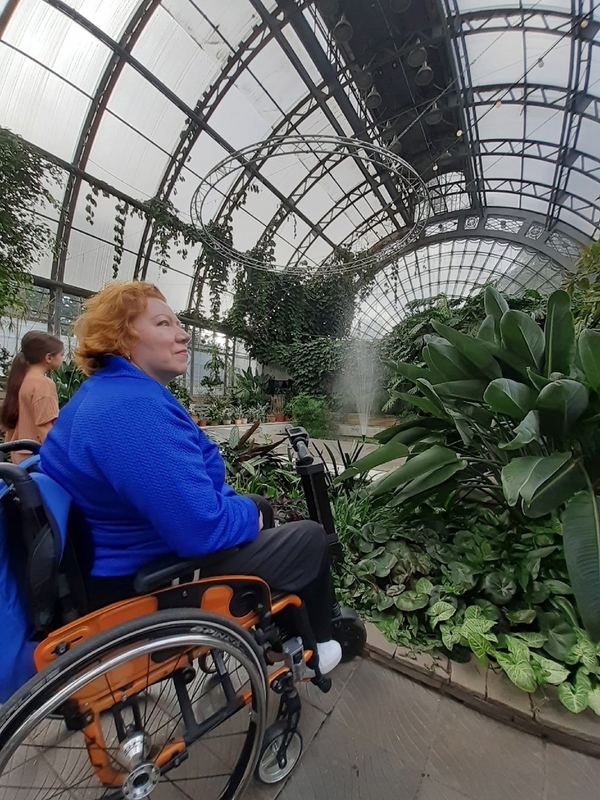 На фото: Марина Козикова на коляске с электроприставкой в оранжерее в окружении тропических растений 