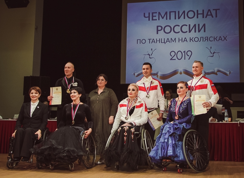 На фото: призеры чемпионата с медалями и грамотами в руках на сцене перед большим экраном на котором написано: "Чемпионат России по танцам на колясках 2019"