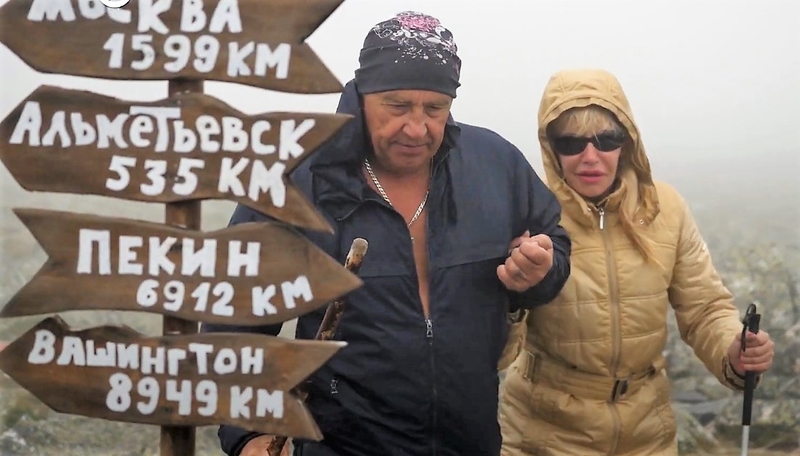 На фото: волонтер - крепкий мужчина в походной одежде, сопровождает Юлию Шумову на маршруте. Туристы идут по тропе в тумане.  Слева от людей стоит деревянный указатель с расстояниями до разных городов мира: Москва, Артемьевск, Пекин, Вашингтон   