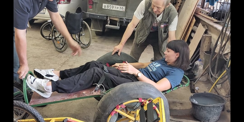 На фото: гараж изобретателей. Девушка в спортивных брюках, футболке и кроссовках полулежит в специальной адаптированной  коляске с большими колесами. Рядом с ней двое мужчин поддерживают коляску. Рядом стоит обычная инвалидная коляска, на полу гаража лежат инструменты, а вдоль стены стоят машины