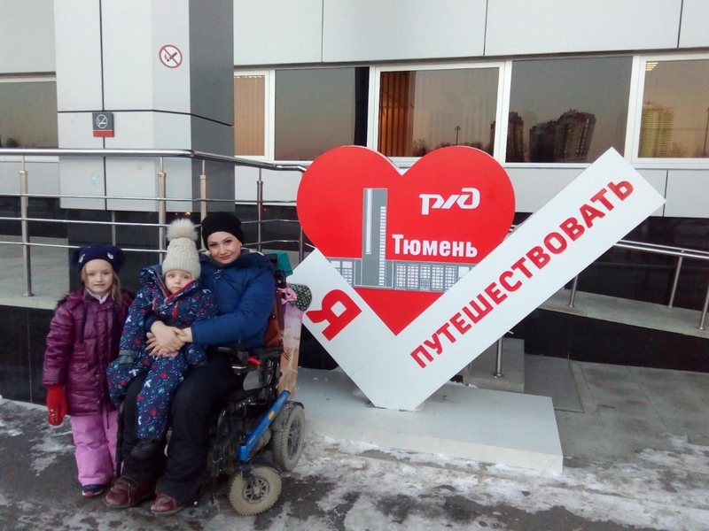 На фото: Ольга Миронова на коляске с детьми возле большой скульптурной композиции на вокзале Тюмени: красное сердце и надпись я люблю путешествовать. Тюмень.