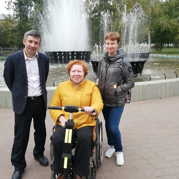 На фото: Марина К - молодая женщина в яркой желтой куртке с рыжими волосами на коляске фотографируется у фонтана на городской площади вместе с двумя гидами - Андреем Даниловым и Дарьей Логуновой