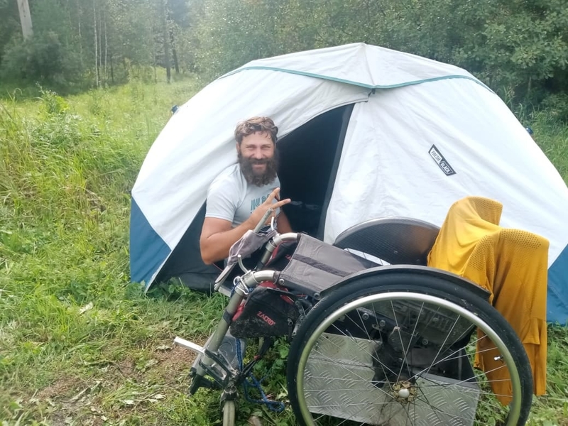 На фото: Виктор Сибирин - мужчина крепкого телосложения с бородой сидит у входа в палатку, рядом с палаткой стоит инвалидная коляска