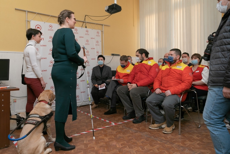 На фото: две девушки – участницы проекта, вместе с собаками-проводниками в специальных шлейках, стоят перед сотрудниками ЦОМП Мосметро, одетыми в форменные красно-желтые куртки и сидящими на стульях в два ряда. Рядом стоит фотограф с висящим на шее большим зеркальным фотоаппаратом. 
