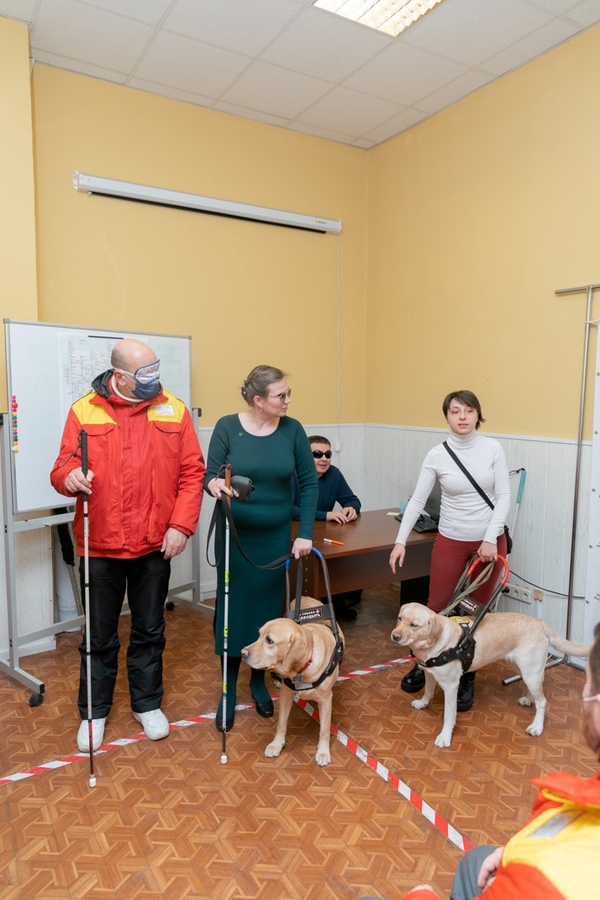 На фото: демонстрация работы с собакой-проводником. Сотрудник ЦОМП в форменной куртке с повязкой на глазах стоит перед аудиторией, держа в правой руке белую трость. Рядом с ним – женщина – ведущая мастер-класса в длинном зеленом платье. В правой руке она держит белую трость, в левой – собаку-проводника за шлейку. Рядом с ней стоит вторая ведущая тоже с собакой-проводником.   