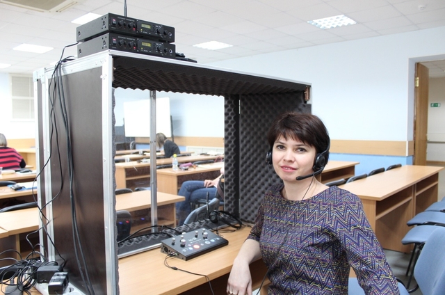 На фото: молодая женщина сидит на стуле у пульта тифлокомментатора с наушниками и микрофоном